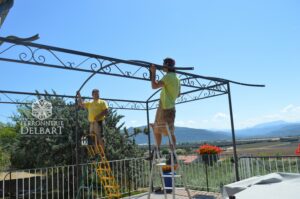 l'équipe de ferronniers positionne une pergola en fer forgé sur mesure sur une terrasse entourée d'un garde-corps acier et d'un arbre sous un ciel bleu ferronnerie delbart création artisanale les Mées Haute Provence (04)